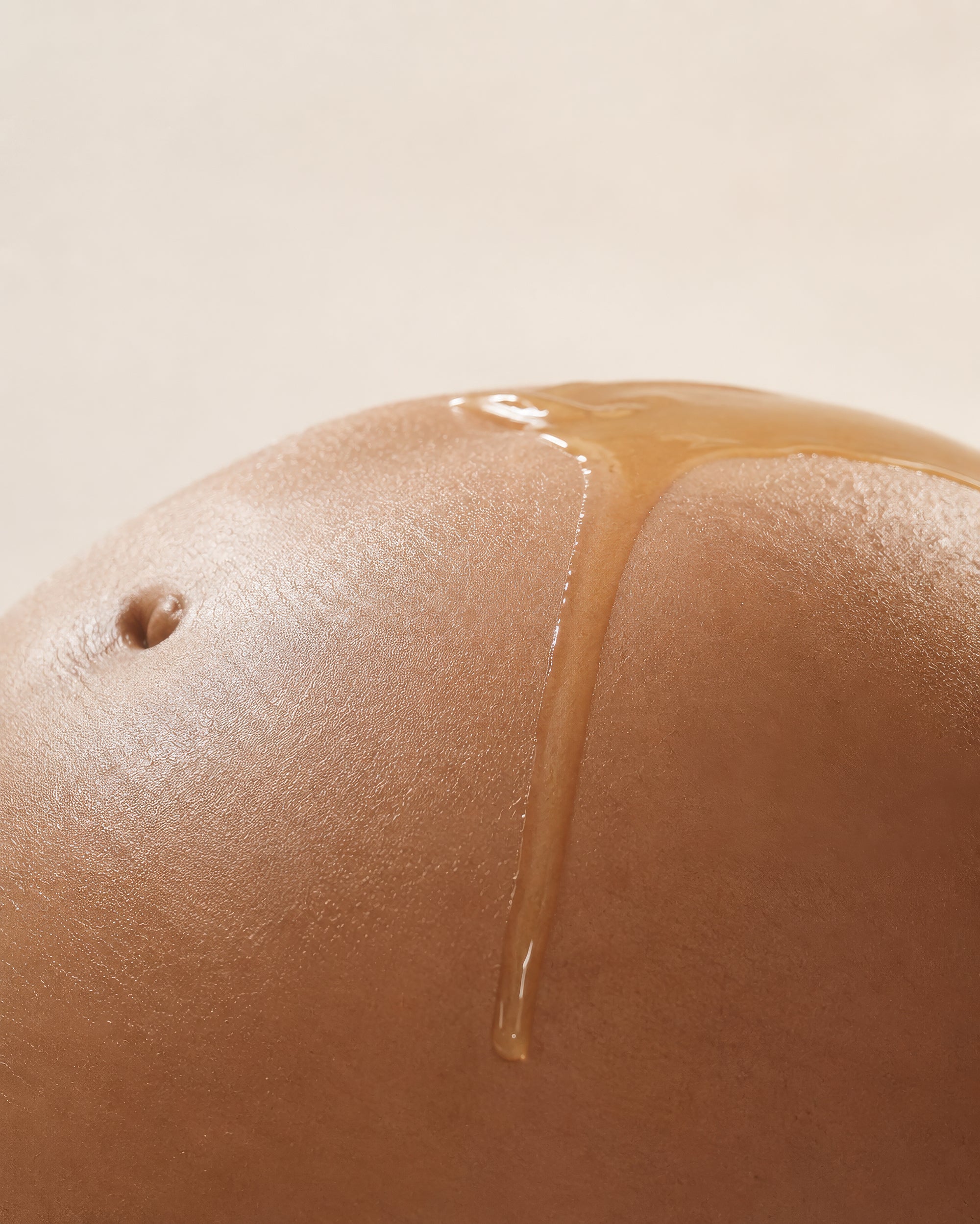 Close-up of a smooth, rounded brown pregnant belly with golden amber liquid belly oil drizzled across its surface against a warm beige background.