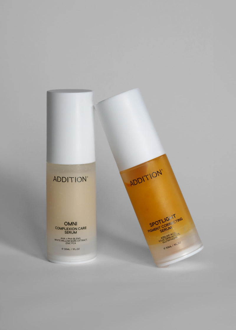 Two ADDITION skincare bottles on a light gray background: a frosted beige serum bottle standing upright and an amber-toned treatment bottle leaning diagonally.