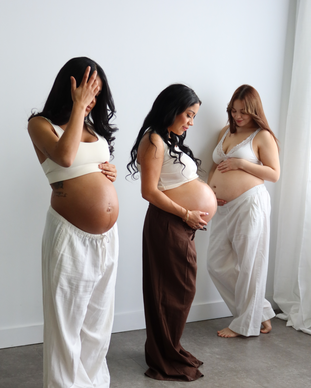 Three pregnant women standing in a studio, wearing white and brown maternity outfits. The woman on the left wears a cream crop top and white pants, the middle woman wears a cream crop top and brown pants, and the woman on the right wears a white crop top and white pants.

